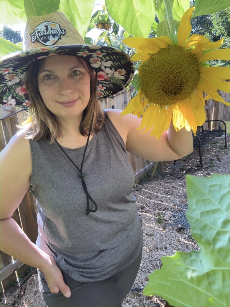 Woman in Farmer's Defense straw hat holding a sunflower in a home garden.