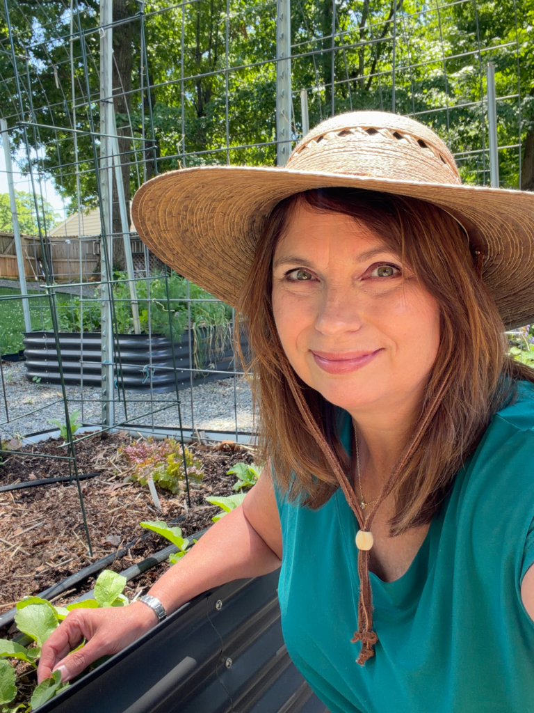 Michelle standing in her backyard garden wearing a green shirt and straw hat beside raised garden beds.